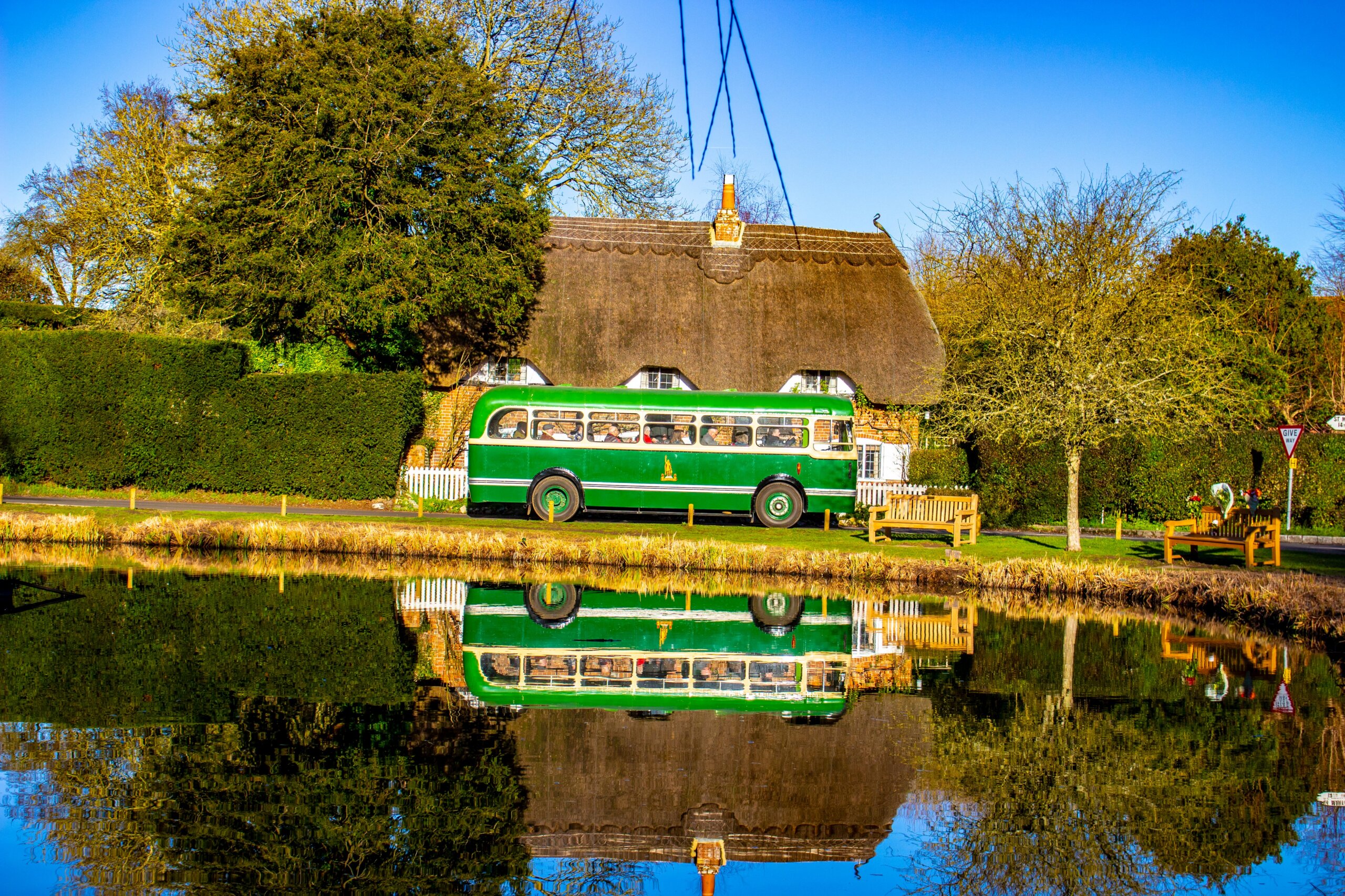 A historic green bus parked in front of a thatch-roofed cottage. The bus and cottage are reflected in the surface of the village's lake.